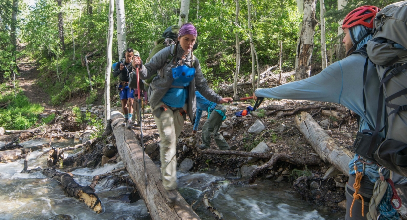 A person crosses a creek over a log while another reaches out their arm to support them. 